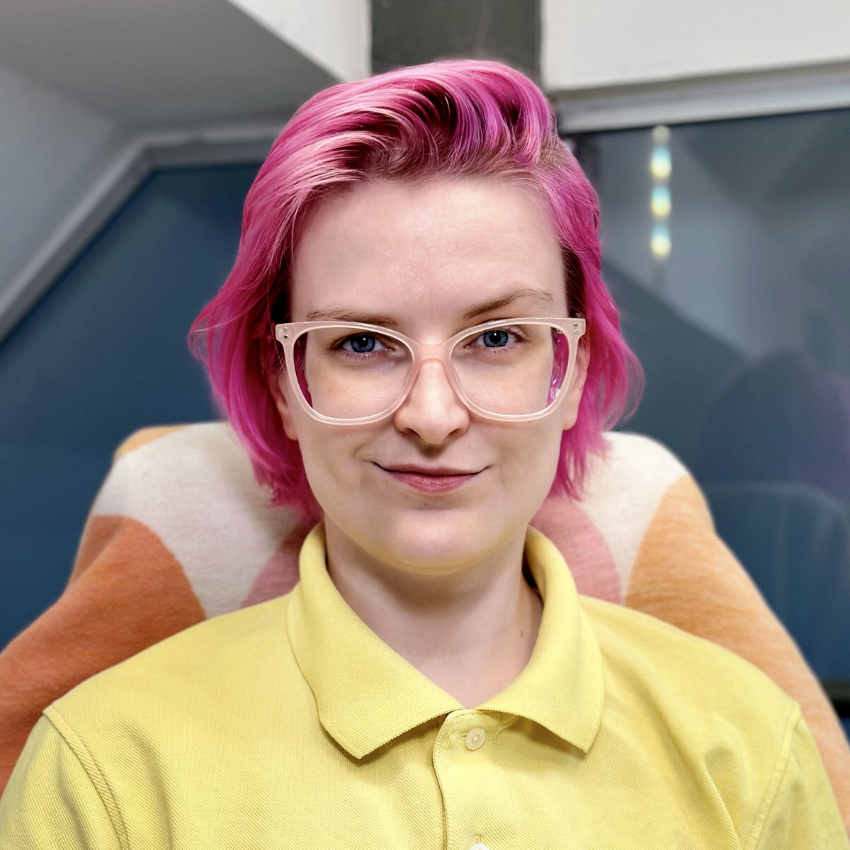 A portrait of Alja Isaković looking directly at the camera with a half smile. Alja has short pink hair, and is wearing light pink glasses and a yellow polo shirt.