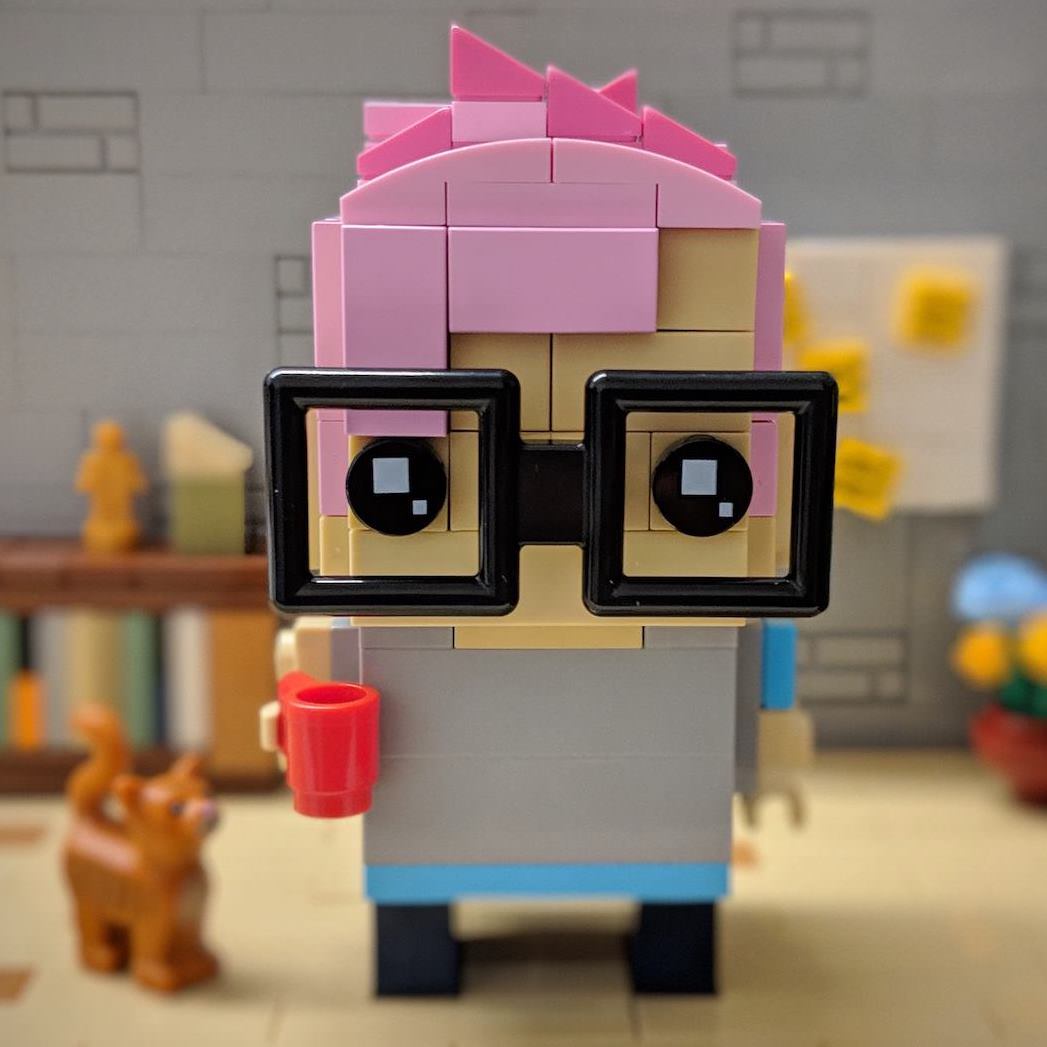 Alja Isaković built with LEGO bricks in the style of LEGO BrickHeadz sets. The brick-built Alja is standing in an office built out of LEGO, with a mug in her hand and an orange LEGO cat looking at the mug.